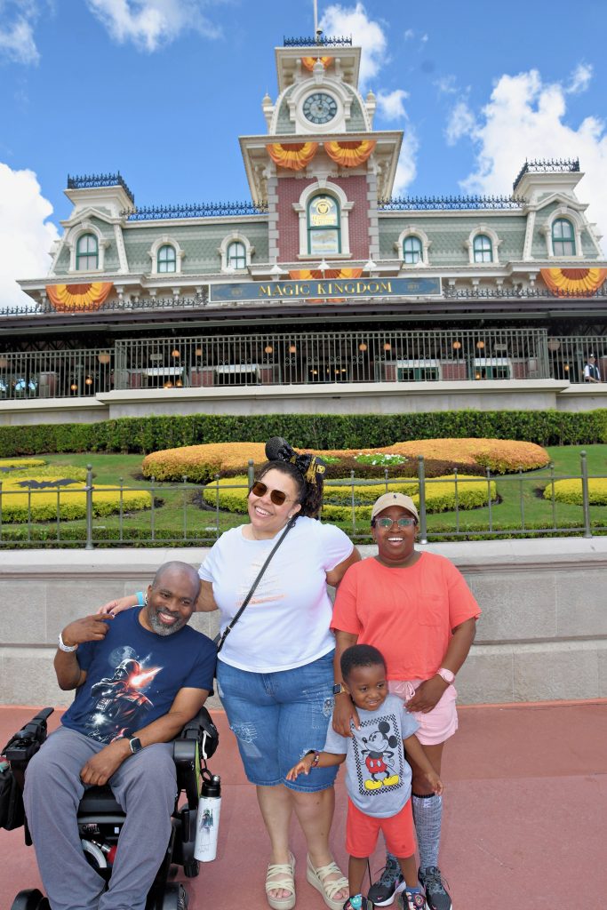 Two women, a man in a wheelchair, and a child pose in front of the entrance to a theme park.